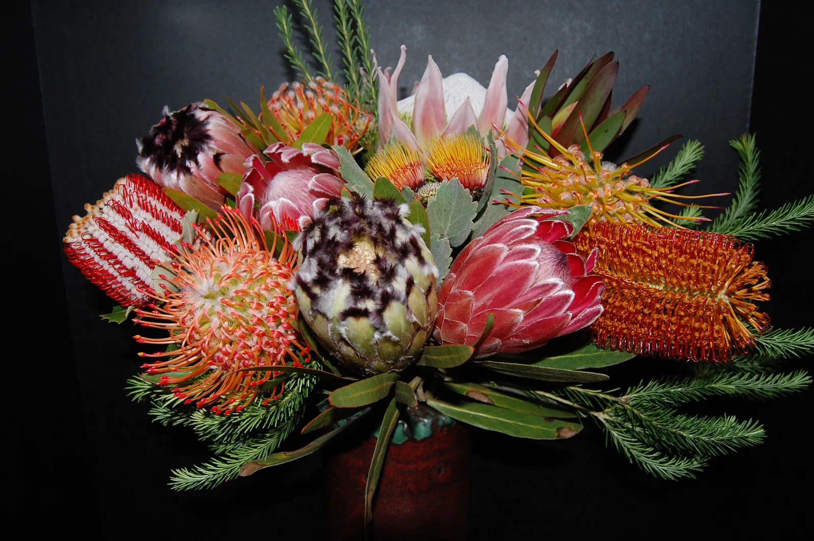 A hand-tied protea arrangement: a pale-pink King protea, deep-pink queen proteas, orange pincushions, dark-tipped mink proteas and red banksia, laced with eucalyptus.